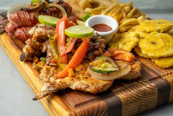 A wooden board featuring a variety of grilled and fried foods including grilled chicken topped with colorful bell peppers and lime, fried plantains, sausages, and a small bowl of dipping sauce.