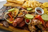 A wooden platter filled with a variety of grilled meats including sausages, steak, and chicken, garnished with lime slices and a colorful mix of bell peppers and onions. Accompanying the meats are fried yuca sticks, French fries, and a small dish of sauce. In the background, there are also fried plantains.