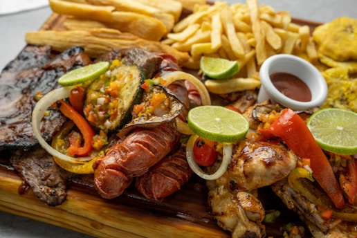 A wooden platter filled with a variety of grilled meats including sausages, steak, and chicken, garnished with lime slices and a colorful mix of bell peppers and onions. Accompanying the meats are fried yuca sticks, French fries, and a small dish of sauce. In the background, there are also fried plantains.