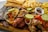 A wooden platter filled with a variety of grilled meats including sausages, steak, and chicken, garnished with lime slices and a colorful mix of bell peppers and onions. Accompanying the meats are fried yuca sticks, French fries, and a small dish of sauce. In the background, there are also fried plantains.