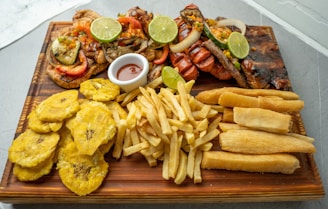 A wooden platter filled with various fried and grilled foods, including grilled meats topped with sliced limes, onions, and peppers. Accompanying the meats are fried plantains, yuca fries, French fries, and a small dish of dipping sauce.