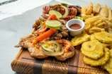 A wooden board is laden with grilled chicken topped with grilled vegetables and lime slices, accompanied by a variety of other foods including fried plantains, sausages, and a serving of fries. A small white dish contains a red sauce for dipping.