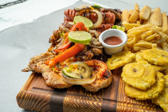 A wooden board is laden with grilled chicken topped with grilled vegetables and lime slices, accompanied by a variety of other foods including fried plantains, sausages, and a serving of fries. A small white dish contains a red sauce for dipping.