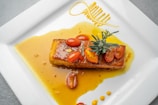 A gourmet dish featuring a glazed piece of fish topped with sliced cherry tomatoes and a sprig of rosemary. The dish is elegantly arranged on a square white plate with a bright yellow sauce artistically drizzled around the fish.