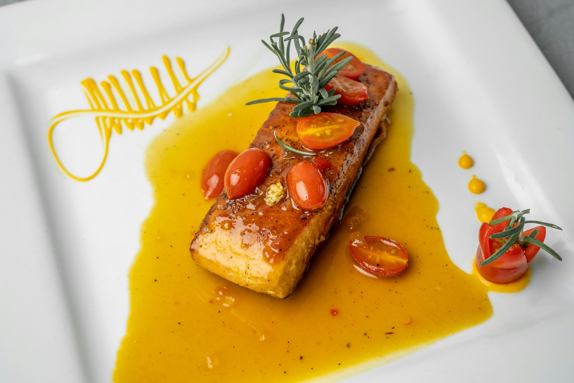 A beautifully presented dish featuring a glazed piece of fish on a white plate, garnished with sprigs of rosemary and halved cherry tomatoes. The fish sits in a pool of vibrant yellow-orange sauce with decorative sauce patterns on the plate.