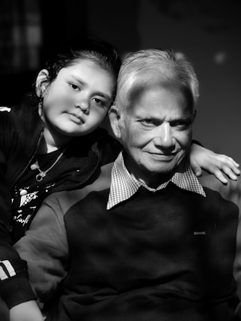 An older man and a young child are closely posed together, both looking directly at the camera. The man wears a dark sweater with a checkered collar, while the child wears a jacket. The light creates a soft, intimate atmosphere, with shadows adding depth to their expressions. The photo is in black and white, enhancing the focus on their facial features.