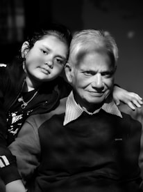 An older man and a young child are closely posed together, both looking directly at the camera. The man wears a dark sweater with a checkered collar, while the child wears a jacket. The light creates a soft, intimate atmosphere, with shadows adding depth to their expressions. The photo is in black and white, enhancing the focus on their facial features.