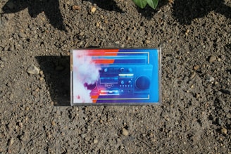 An audio cassette tape with a cover art featuring a retro-style boombox design placed on a textured, earthy ground. The cover art is vivid with a mix of blue, red, and white colors, accentuating the boombox imagery.