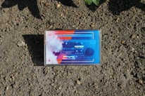 An audio cassette tape with a cover art featuring a retro-style boombox design placed on a textured, earthy ground. The cover art is vivid with a mix of blue, red, and white colors, accentuating the boombox imagery.