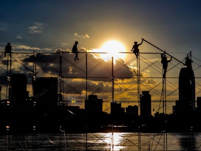 Close-up of hands wearing work gloves holding a steel beam with a city skyline in the distance during sunset.