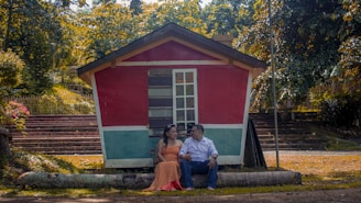 A couple sitting on a log in front of a small, colorful wooden house with red and teal walls, surrounded by lush greenery. They are holding hands and gazing at each other, creating a serene and romantic atmosphere.