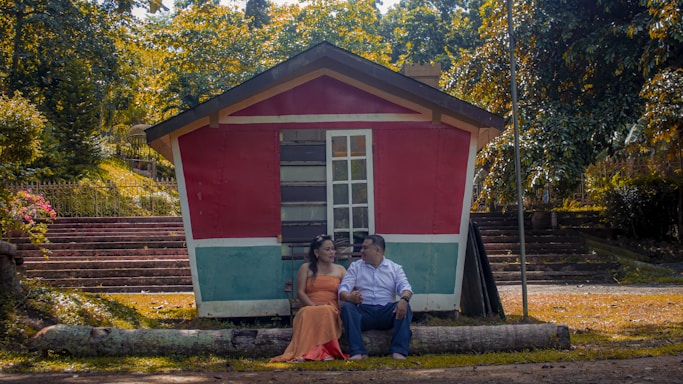 A couple sitting on a log in front of a small, colorful wooden house with red and teal walls, surrounded by lush greenery. They are holding hands and gazing at each other, creating a serene and romantic atmosphere.