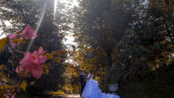 A couple stands together in a lush garden setting, with the sun shining brightly through the tree canopy, creating a romantic atmosphere. The foreground features vibrant pink flowers while large trees and greenery surround the couple, adding depth and serenity to the scene.