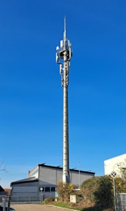 A tall communication tower stands prominently in front of a white and gray industrial building. The tower is equipped with various antennas and transmitters. The sky is clear and blue, and there is a small grassy area with bushes and a street sign in the foreground. The scene is well-lit by sunlight, casting minimal shadows.