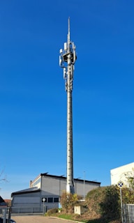 A tall communication tower stands prominently in front of a white and gray industrial building. The tower is equipped with various antennas and transmitters. The sky is clear and blue, and there is a small grassy area with bushes and a street sign in the foreground. The scene is well-lit by sunlight, casting minimal shadows.
