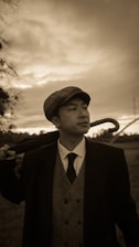 A man dressed in vintage-style clothing, including a flat cap, vest, tie, and long coat, stands outdoors holding a cane over his shoulder. The sky behind him is overcast, casting a sepia tone over the scene.