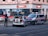 A city street scene with police officers outside a store named 'tipwin'. Two police vehicles, a car and a van, are parked on the road. One officer stands near the police car with its trunk open, while another stands beside the van. The environment suggests a possibly serious or tense situation.