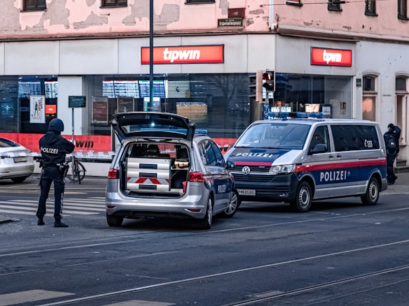 A city street scene with police officers outside a store named 'tipwin'. Two police vehicles, a car and a van, are parked on the road. One officer stands near the police car with its trunk open, while another stands beside the van. The environment suggests a possibly serious or tense situation.