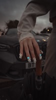 A hand with intricate nail art featuring blue and white designs rests on a wooden umbrella handle. The background includes a dark-colored car with the license plate partially visible. The overall setting is dimly lit, suggesting evening or overcast weather.
