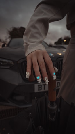 A hand with intricate nail art featuring blue and white designs rests on a wooden umbrella handle. The background includes a dark-colored car with the license plate partially visible. The overall setting is dimly lit, suggesting evening or overcast weather.