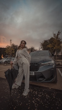 A person stands confidently in front of a dark grey car, holding an umbrella. They are wearing a light-colored suit and white sneakers. The sky above is overcast, hinting at an impending storm, and the surrounding trees frame the parking lot scene.