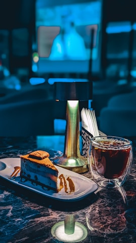 A slice of layered cake garnished with caramel drizzle sits on a rectangular white plate. Next to it is a glass cup filled with dark coffee. These items are placed on a marble countertop with a small, modern, gold-toned lamp in between, adding a warm glow. In the background, there appears to be a blurry projection of an image, suggesting a cozy cafe or lounge setting.
