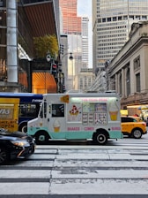 An ice cream truck is parked at a pedestrian crosswalk in a busy city environment, surrounded by large buildings and a yellow taxi. The truck has colorful illustrations of ice cream cones, shakes, and sundaes. There are other vehicles, including a black car and a hybrid bus, nearby. The street is bustling with activity.