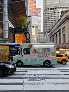 An ice cream truck is parked at a pedestrian crosswalk in a busy city environment, surrounded by large buildings and a yellow taxi. The truck has colorful illustrations of ice cream cones, shakes, and sundaes. There are other vehicles, including a black car and a hybrid bus, nearby. 