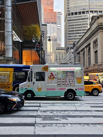 An ice cream truck is parked at a pedestrian crosswalk in a busy city environment, surrounded by large buildings and a yellow taxi. The truck has colorful illustrations of ice cream cones, shakes, and sundaes. There are other vehicles, including a black car and a hybrid bus, nearby. The street is bustling with activity.