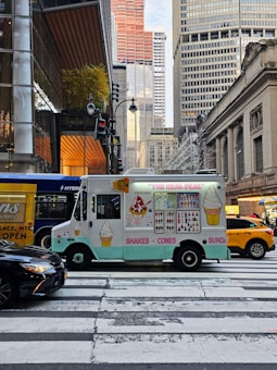 An ice cream truck is parked at a pedestrian crosswalk in a busy city environment, surrounded by large buildings and a yellow taxi. The truck has colorful illustrations of ice cream cones, shakes, and sundaes. There are other vehicles, including a black car and a hybrid bus, nearby. The street is bustling with activity.