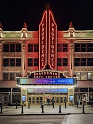 The image features the front facade of a performing arts center named Providence. The building is ornately decorated, with a large vertical neon sign displaying 'PROVIDENCE' twice. The entrance is brightly lit with a marquee sign advertising a '2024 IPMA launch party' scheduled for December 9, 2023. The architecture is detailed and well-lit, especially in the nighttime setting. There are two people standing near the entrance.