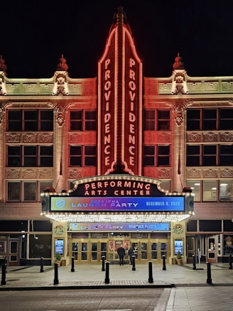 The image features the front facade of a performing arts center named Providence. The building is ornately decorated, with a large vertical neon sign displaying 'PROVIDENCE' twice. The entrance is brightly lit with a marquee sign advertising a '2024 IPMA launch party' scheduled for December 9, 2023. The architecture is detailed and well-lit, especially in the nighttime setting. There are two people standing near the entrance.