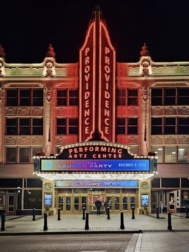 The image features the front facade of a performing arts center named Providence. The building is ornately decorated, with a large vertical neon sign displaying 'PROVIDENCE' twice. The entrance is brightly lit with a marquee sign advertising a '2024 IPMA launch party' scheduled for December 9, 2023. The architecture is detailed and well-lit, especially in the nighttime setting. There are two people standing near the entrance.