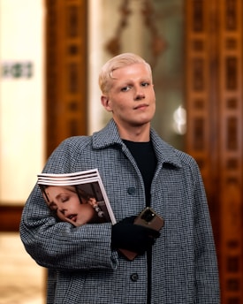 A person with short platinum blonde hair is dressed in a stylish plaid coat and is holding a magazine and a smartphone. The setting appears to be indoors, with ornate wooden paneling visible in the background.