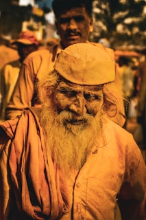 A group of people dressed in earthy, orange-toned garments is captured. The focus is on an elder man with a long beard, wearing a cap and a traditional cotton robe. Behind him, a man stands in the blurred background. The vibrant lighting accentuates the textures in their clothing and skin, casting a warm glow across the scene.