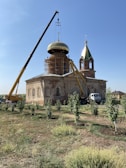 A construction site featuring a church with two cranes lifting a golden dome into place. The building appears to be old, with ornate architectural details. Scaffolding surrounds part of it, and there are small trees and grass in the foreground.