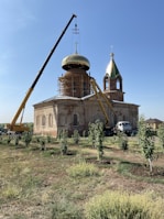 A construction site featuring a church with two cranes lifting a golden dome into place. The building appears to be old, with ornate architectural details. Scaffolding surrounds part of it, and there are small trees and grass in the foreground.