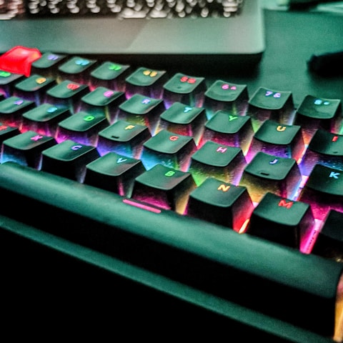 An RGB mechanical keyboard with black keycaps illuminated by vibrant multicolored backlighting. The lights create a rainbow effect, highlighting the keys with various colors such as red, blue, green, and yellow. A red escape key stands out prominently, and the keyboard is placed on a dark surface.