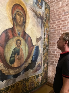 A man stands closely observing an intricately painted religious icon featuring a central figure with a halo, possibly depicting a biblical scene. The icon is set on a textured brick background.