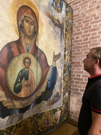 A man stands closely observing an intricately painted religious icon featuring a central figure with a halo, possibly depicting a biblical scene. The icon is set on a textured brick background.