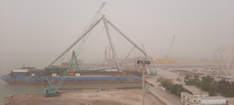 A large construction site features multiple cranes and a large ship docked at a shipyard. The scene is hazy, likely due to mist or pollution, and several industrial buildings and equipment are visible. The ground is dusty, and there are trees and structures along the right side.