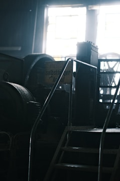 A dimly lit industrial setting featuring metal stairs leading up to machinery. A sign with German text is visible, partially obscured by shadows. The background shows diffused light entering through a window.