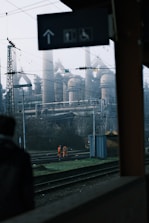 Workers in orange safety clothing walk along train tracks in an industrial setting. In the background, large industrial structures with smokestacks are visible under a cloudy sky. Overhead cables and signs for pedestrian and wheelchair access are also prominent.