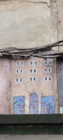 A textured wall art piece depicting a stylized, simple illustration of a rectangular building facade. The building has two columns of windows painted in white squares on a brown-colored surface. At the base of the building, there are blue doors with abstract shapes resembling people. Above the art, there are twigs or wires running horizontally.