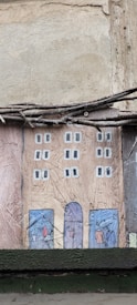 A textured wall art piece depicting a stylized, simple illustration of a rectangular building facade. The building has two columns of windows painted in white squares on a brown-colored surface. At the base of the building, there are blue doors with abstract shapes resembling people. Above the art, there are twigs or wires running horizontally.