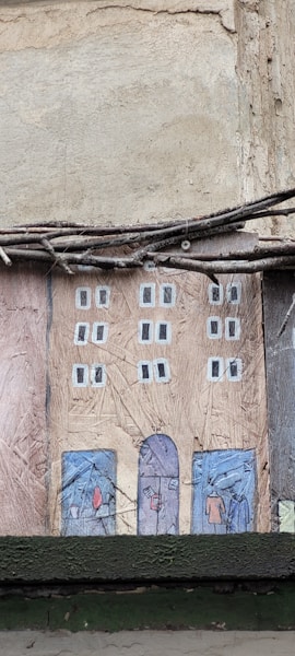 A textured wall art piece depicting a stylized, simple illustration of a rectangular building facade. The building has two columns of windows painted in white squares on a brown-colored surface. At the base of the building, there are blue doors with abstract shapes resembling people. Above the art, there are twigs or wires running horizontally.