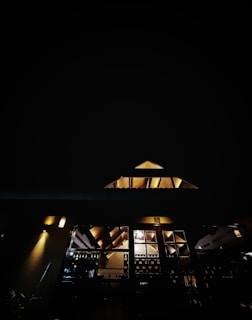 A building with illuminated interior lights is captured against a pitch-dark night sky. The structure has an angular roof design, and several windows are visible with light emanating from within. Light fixtures on the exterior walls also cast a soft glow, highlighting parts of the architectural elements.