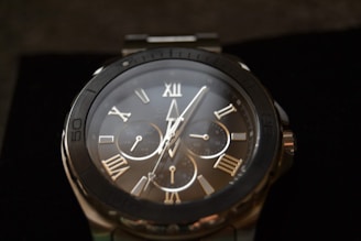 A close-up view of a wristwatch with a metal band. The watch face features Roman numerals and multiple smaller dials, likely indicating various time-related functions. The hands of the watch are pointed and sleek, and the overall style suggests a blend of classic and modern design.