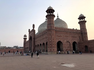 A large, historic mosque with intricate architecture featuring tall minarets and a distinctive large central dome. The walls and arches are adorned with detailed geometric designs. Several people are walking around or sitting on the spacious courtyard made of stone tiles, giving a sense of both grandeur and daily life.