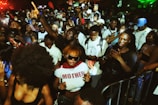 A vibrant crowd of young people enjoying a lively outdoor event. Many are holding smartphones and some are dancing, while others are engaging in conversations. There are bright lights and a dynamic atmosphere with energetic expressions.
